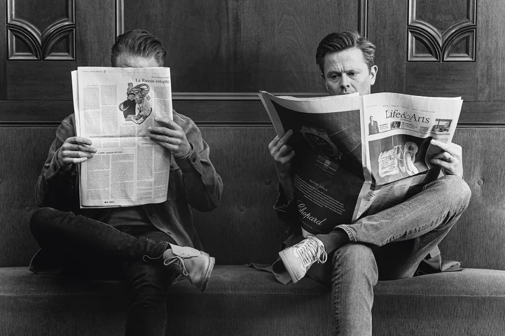 Filip och Fredrik sitter på en bänk och läser varsin tidning. Den ena mannens ansikte är dolt bakom tidningen, medan den andras ansikte syns när han tittar i tidningen. Bilden är i svartvitt.