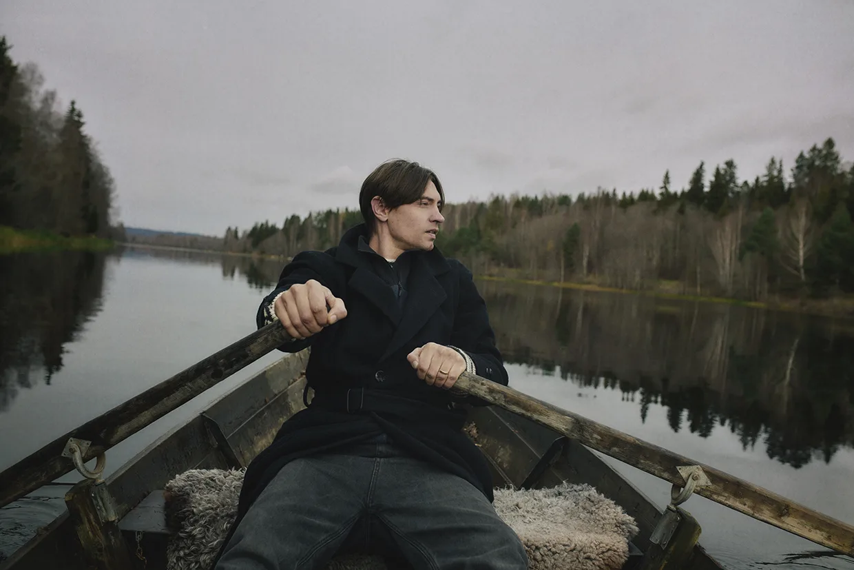 Gustaf Norén i mörk kappa sitter i en roddbåt på en lugn sjö och håller i årorna. Han tittar åt sidan, omgiven av stilla vatten och träd med kala grenar en molnig dag.