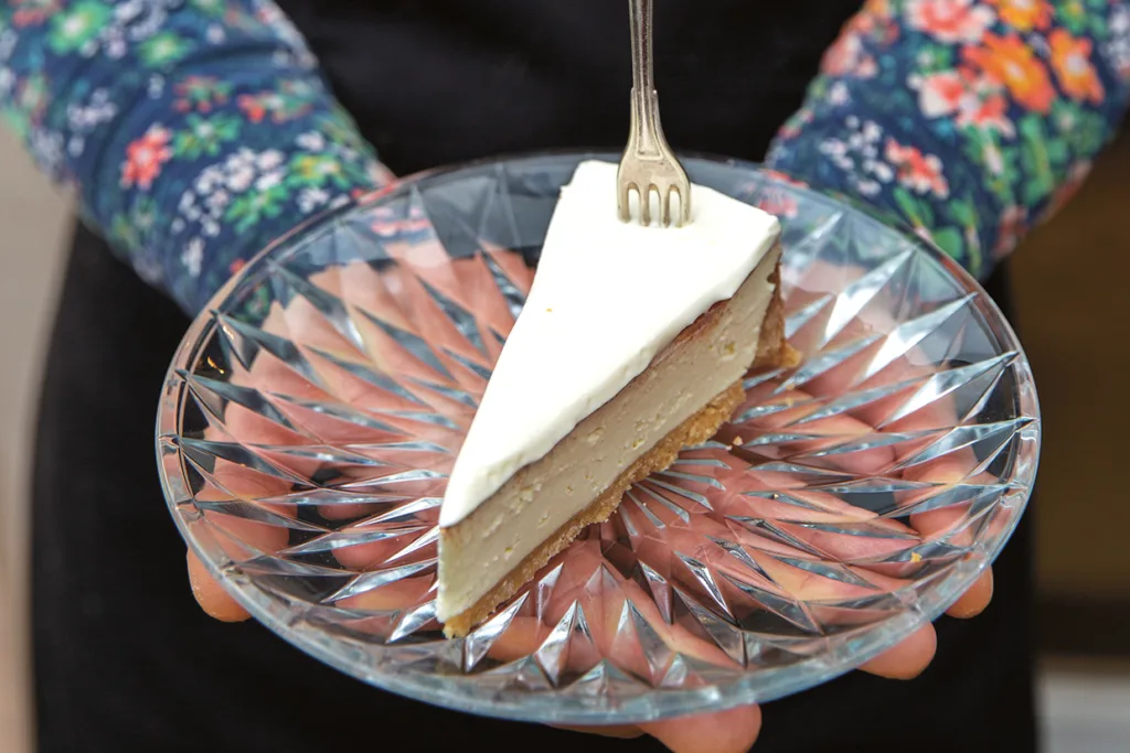En person håller i en klar, mönstrad glastallrik med en skiva cheesecake toppad med ett lager vit grädde och en liten gaffel som sticker in i toppen. Personen är klädd i en blommönstrad långärmad skjorta.