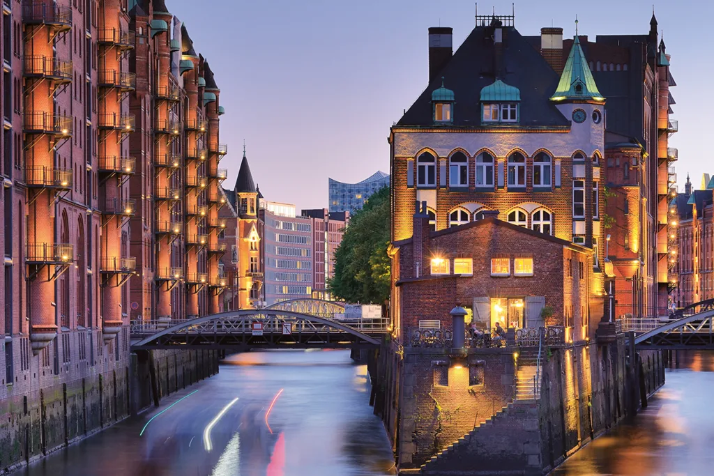 En kanal i skymningen i Hamburgs Speicherstadt-distrikt, med upplysta lagerbyggnader i tegel och en historisk byggnad på en liten ö, som speglar sig i det lugna vattnet.