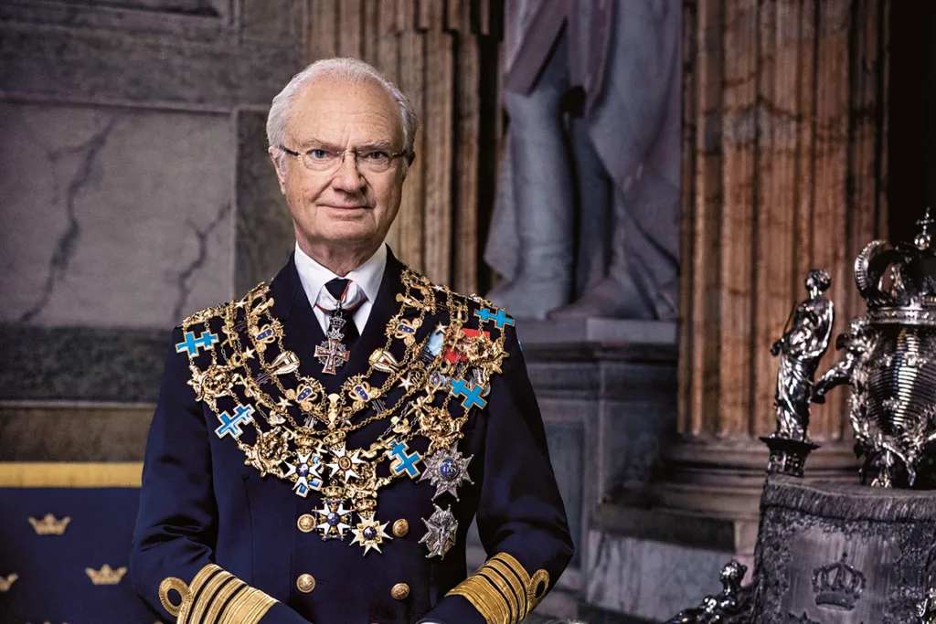 H.M. Konungen med glasögon och en mörk formell uniform prydd med många guldmedaljer, kedjor och kors står i ett utsmyckat rum med statyer och detaljerad arkitektur i bakgrunden.