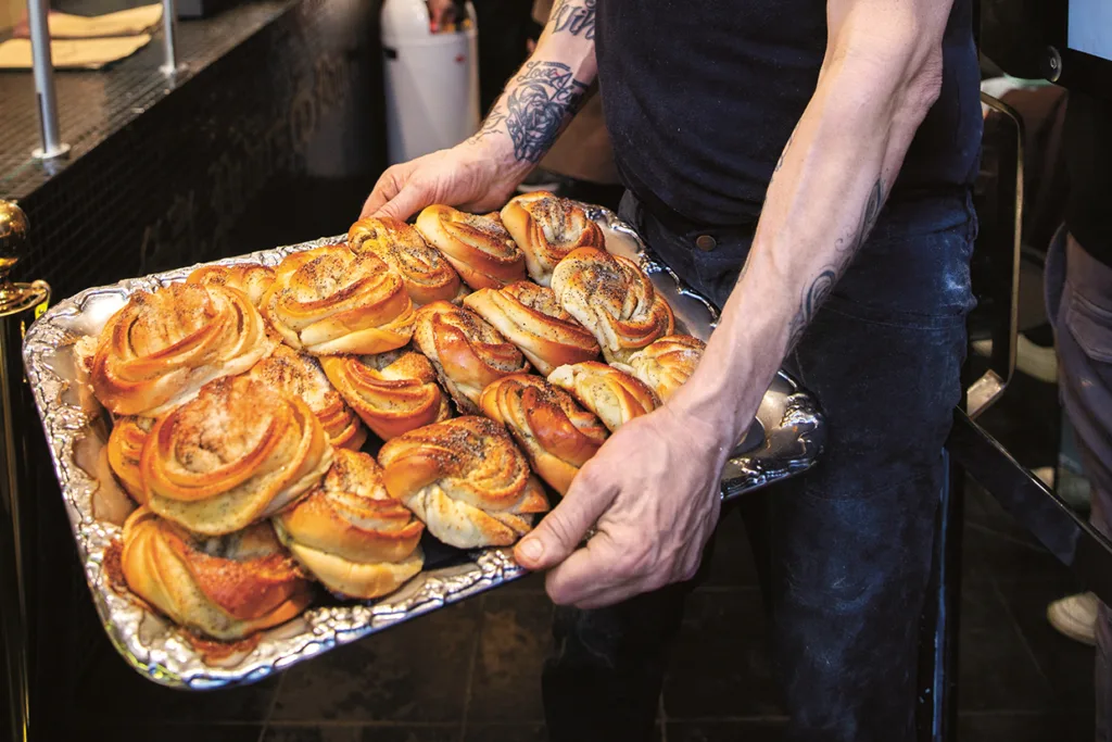 En person med tatuerade armar håller en stor bricka fylld med nygräddade, gyllenbruna kanelbullar i en bagerimiljö.