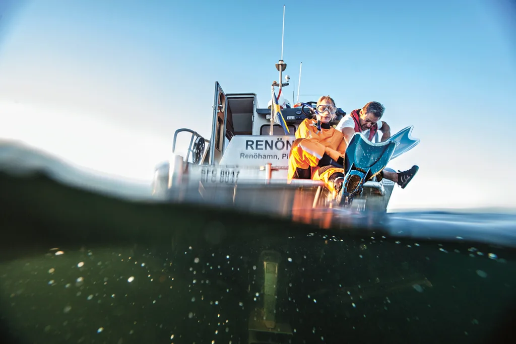 Två personer sitter på kanten av en båt. Den ena har på sig en orange våtdräkt och en snorkelmask och håller i en blå sjöjungfrusvans och är på väg ner i vattnet. Bilden är tagen till hälften ovanför och till hälften under vattenytan.