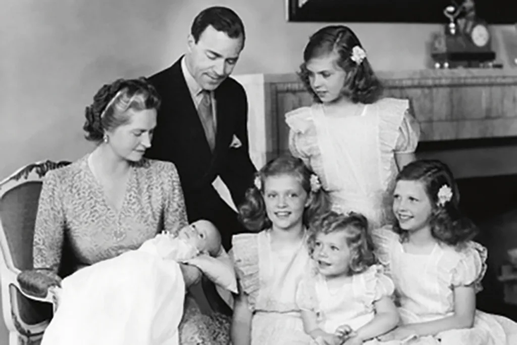 Ett svartvitt foto av kungafamiljen: en sittande kvinna med en bebis, Kungen, i famnen, en man som står bakom henne och fyra unga flickor i matchande klänningar, alla leende, i ett vardagsrum med en öppen spis i bakgrunden.