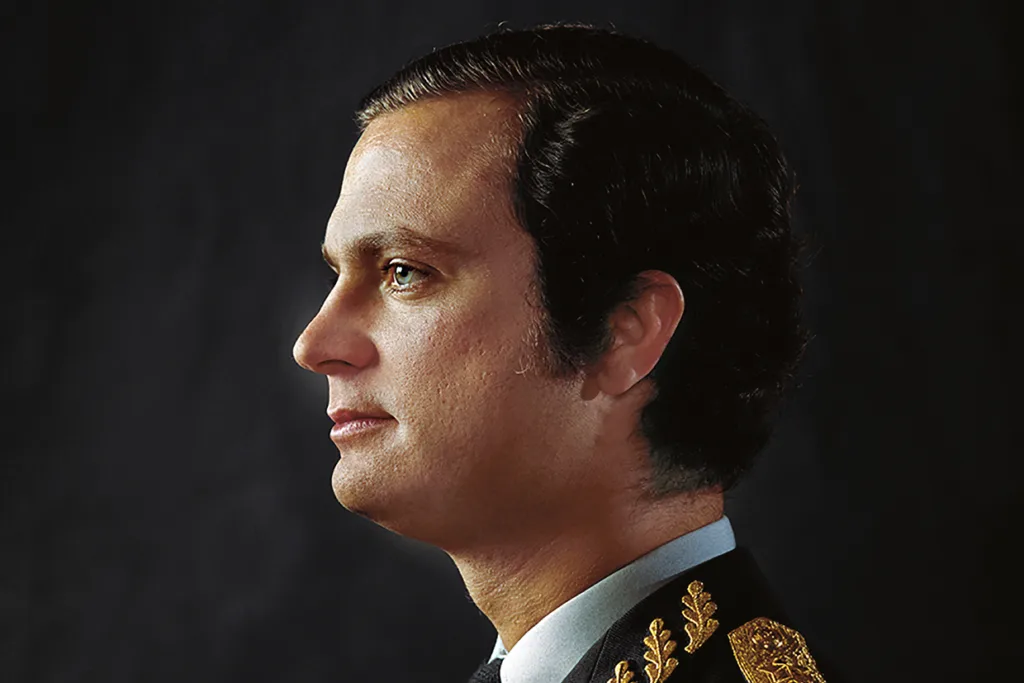 Kungen med mörkt hår i en formell uniform med guldbroderier visas i profil mot en mörk bakgrund.