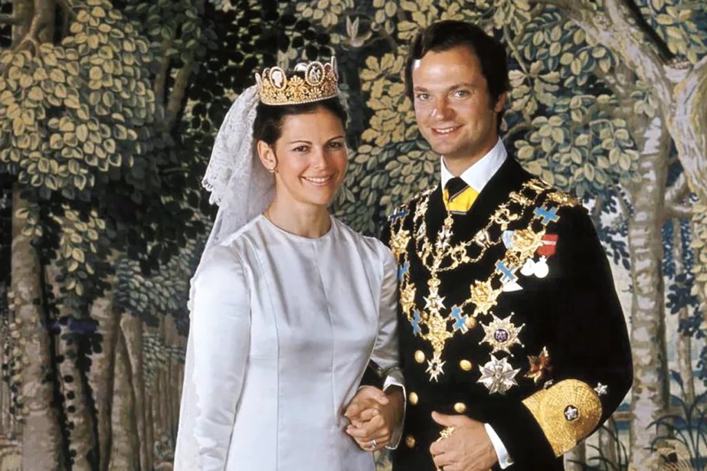 Silvia, Drottningen, i vit klänning och slöja står bredvid Kungen i dekorerad militäruniform, båda ler och håller varandra i handen, med en bakgrund med skogstema bakom dem.