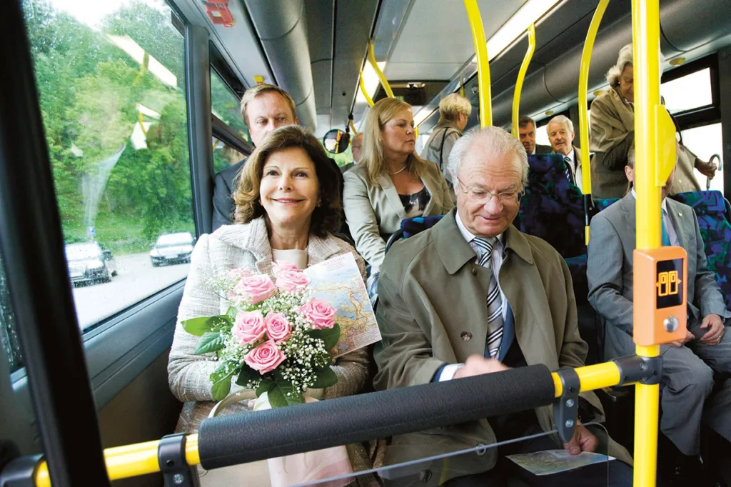 Drottningen håller i en bukett rosa rosor sitter bredvid Kungen i kostym och trenchcoat på en buss, omgiven av andra välklädda passagerare. Stämningen är formell och glad.