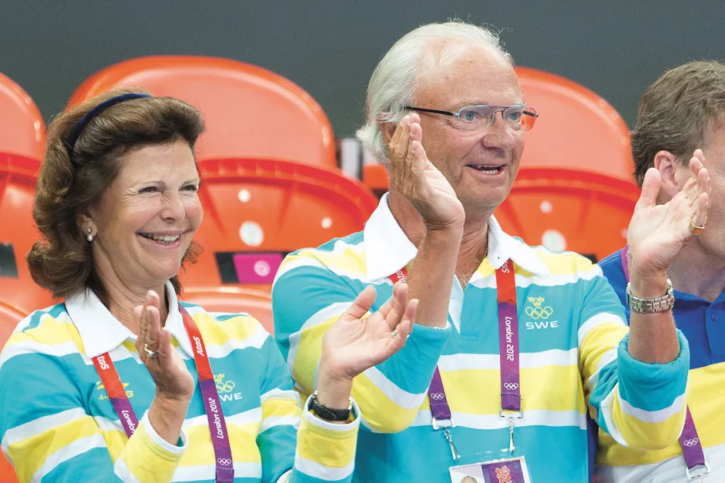 Kungaparet i matchande blå-, gul- och vitrandiga skjortor applåderar på en stadion med orangea säten. Båda har nyckelband runt halsen och ser ut att njuta av ett evenemang.