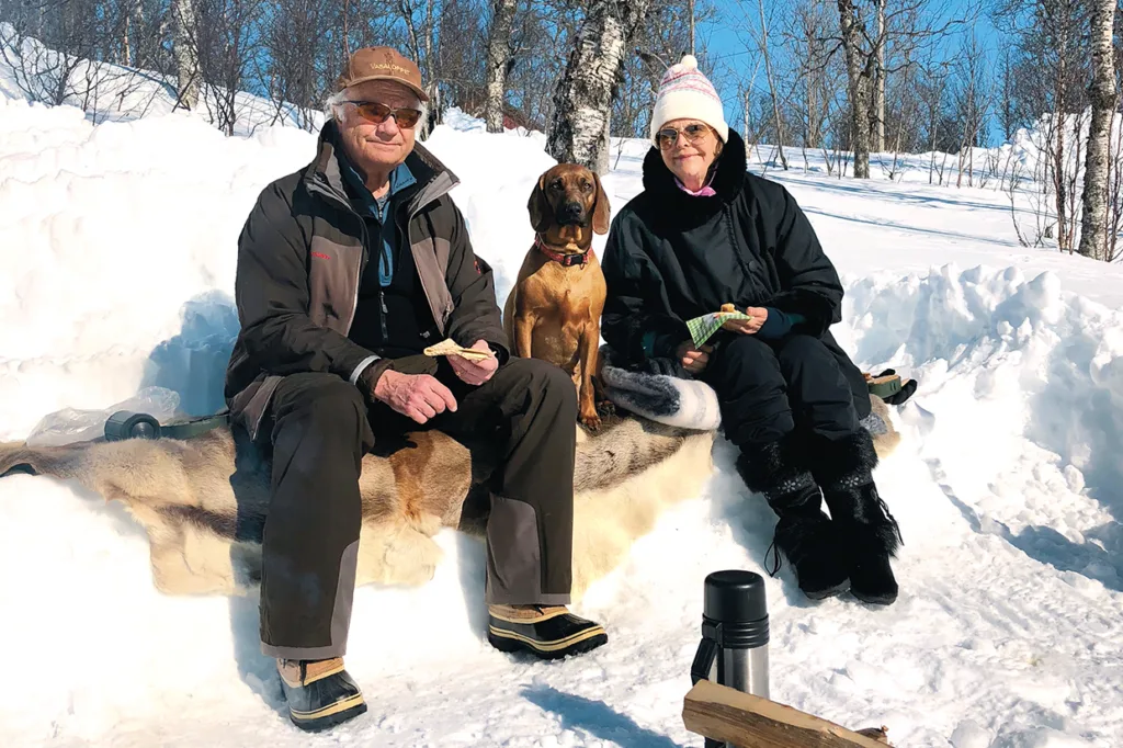 Kungaparet sitter på en bänk täckt av djurpäls i snöig utomhusmiljö, med en brun hund mellan sig. De bär vinterkläder och solglasögon och håller i smörgåsar. En termos och en stock ligger i snön i närheten.