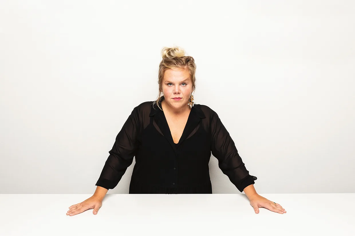 Linnea Wikblad med blont hår i en rufsig knut, klädd i en svart genomskinlig blus, står bakom ett vitt bord med båda händerna vilande på bordet och tittar rakt in i kameran mot en vit bakgrund.