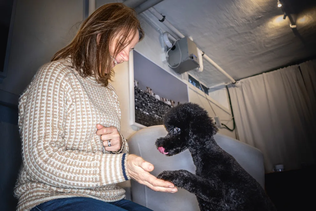 Kicki Fellstenius i beige tröja ler medan hon håller ut handen mot en svart pudel som ger henne sin tass. Hunden tittar på henne och slickar sig om läpparna i en inomhusmiljö.