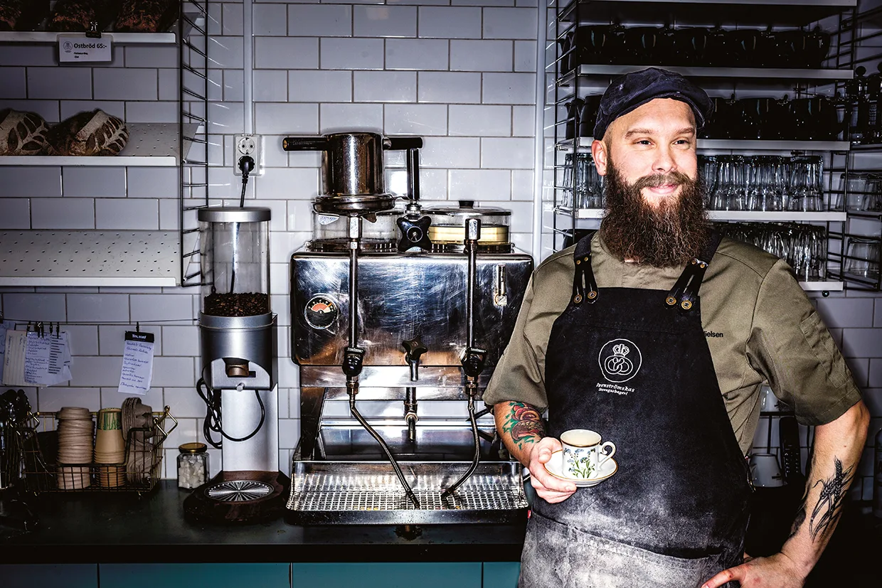 En skäggig man i förkläde står i ett kafékök och håller i en kopp och ett fat, med en espressomaskin och hyllor med koppar och tillbehör bakom sig.
