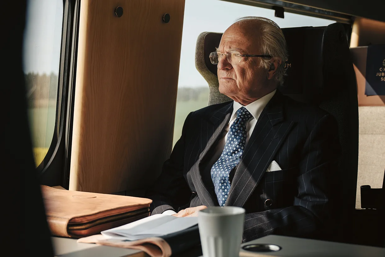 Kungen i kostym och slips sitter vid ett fönster på ett tåg och tittar eftertänksamt ut. På bordet framför honom ligger en mapp, papper och en vit kaffekopp. Solljuset flödar in genom fönstret.