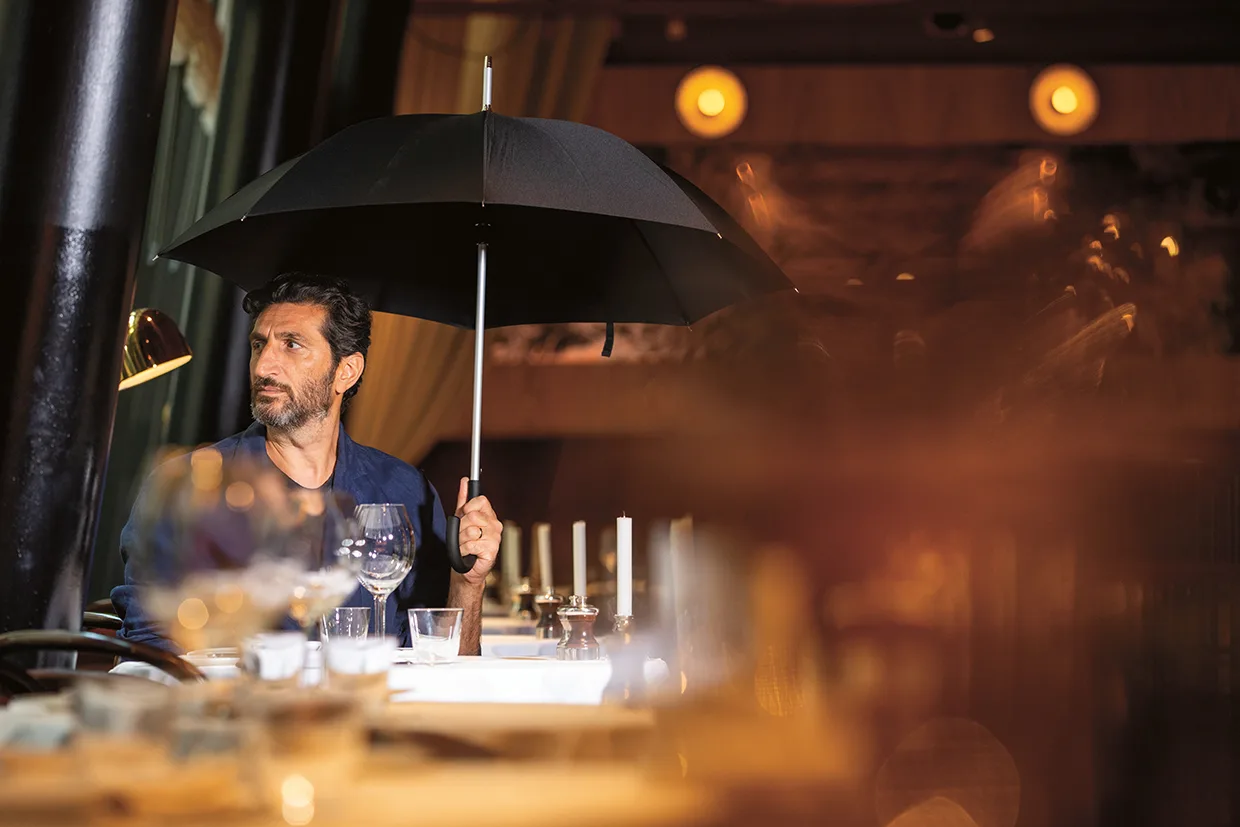 En man sitter ensam vid ett restaurangbord och håller ett öppet svart paraply inomhus, omgiven av tomma glas och tallrikar, med varm belysning i bakgrunden.