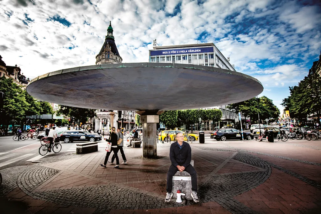 En person sitter på ett betongblock under en stor cirkelformad byggnad på ett livligt torg i en stad, omgiven av fotgängare, cyklister och bilar. I bakgrunden syns byggnader och en delvis molnig himmel.