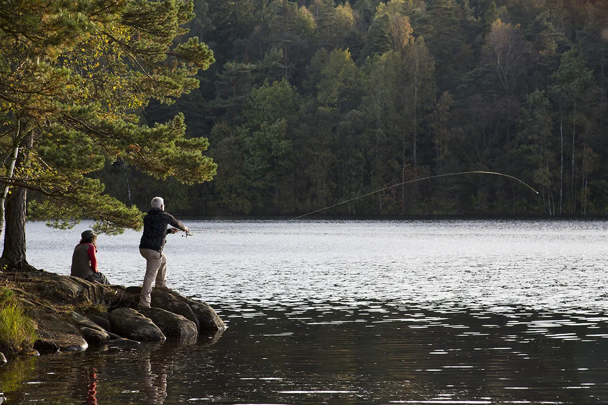 Två personer befinner sig på en klippig strand vid en lugn sjö omgiven av träd. Den ena personen sitter och den andra står och kastar en fiskelina i vattnet. Scenen verkar fridfull och naturlig.