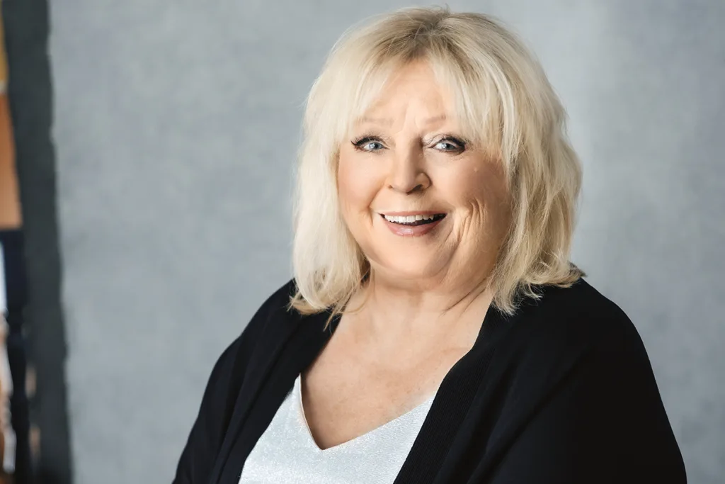 En leende Kicki Danielsson med axellångt blont hår, klädd i en vit topp och en svart kofta, poserar framför en neutral grå bakgrund.