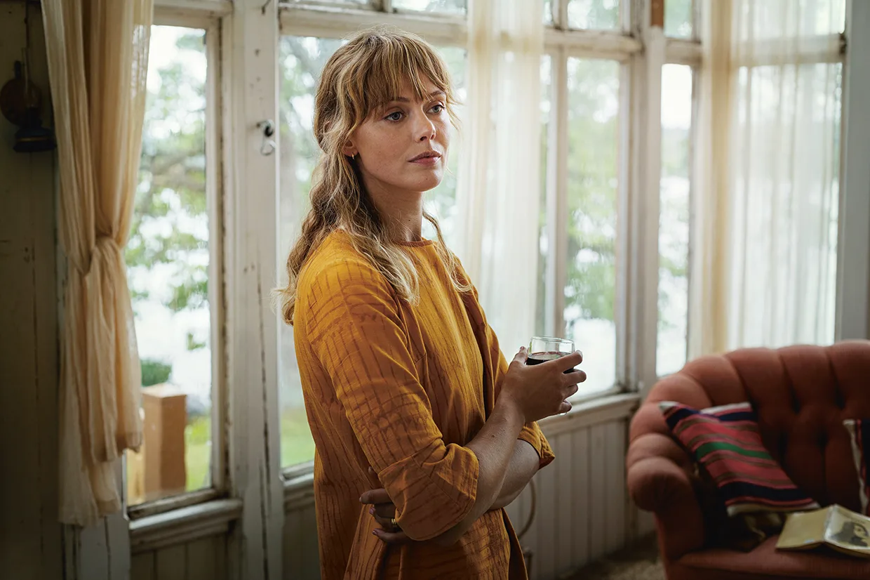 Frida Gustavsson i orange klänning står inomhus vid ett stort fönster med ett glas vatten i handen och ser tankfull ut. Rummet har skira gardiner, en röd soffa och solljus som flödar in.