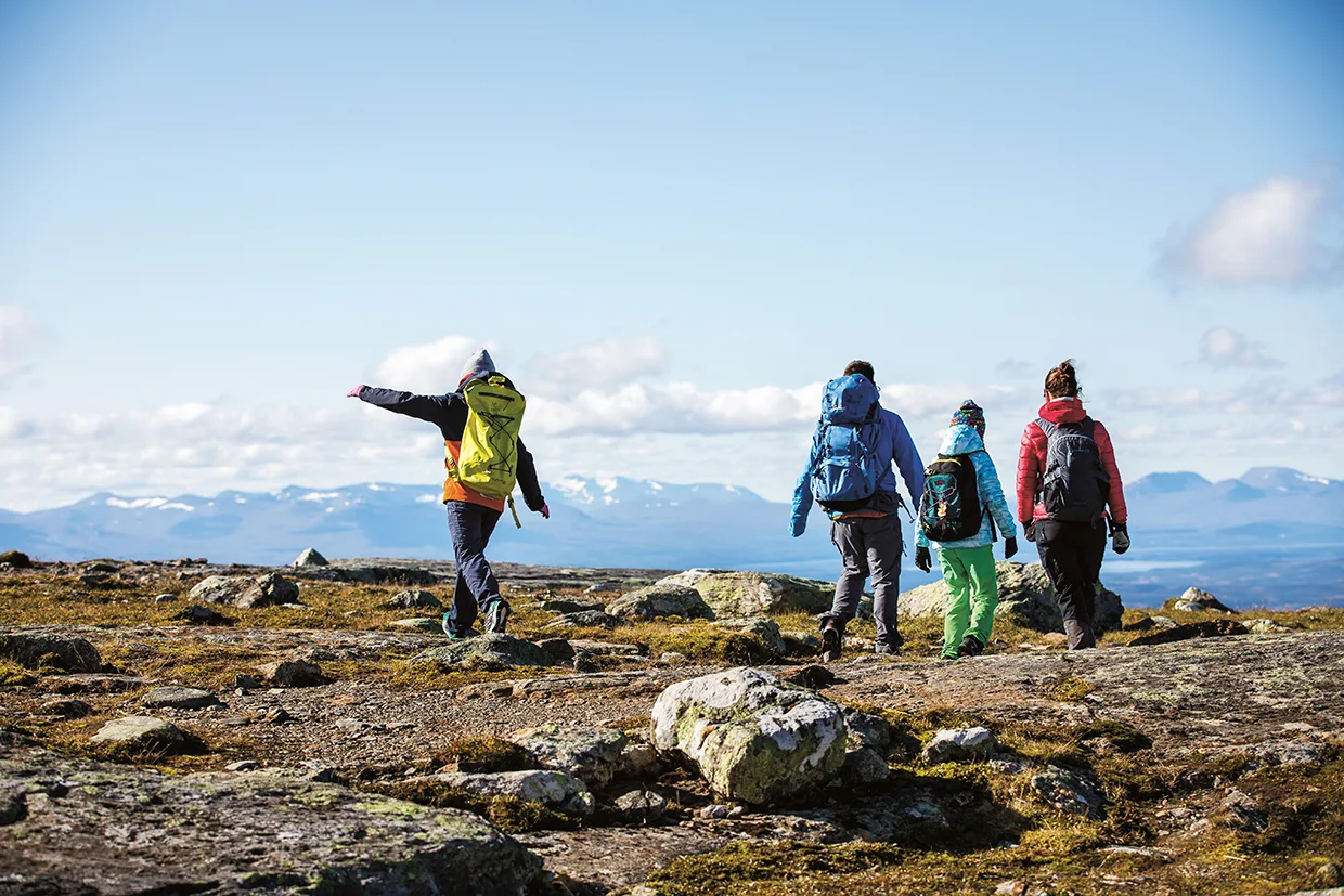 Fyra personer med ryggsäckar vandrar genom ett stenigt, öppet landskap med avlägsna berg och blå himmel i bakgrunden. En person i gul jacka har armarna utsträckta medan han går.