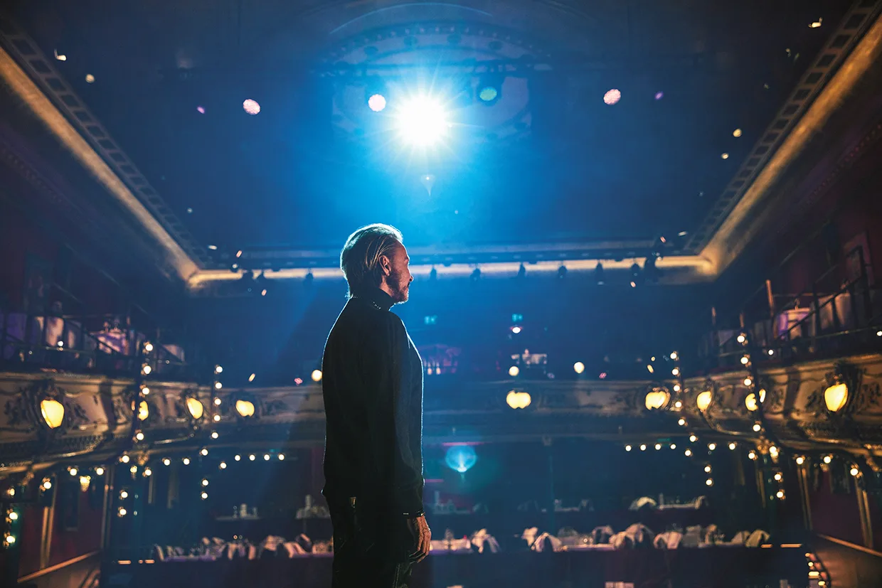 Christer Lindarv står på en teaterscen vänd mot publiken, upplyst av en stark strålkastare, med utsmyckade balkonger och tomma stolar synliga i den svagt upplysta hörsalen.