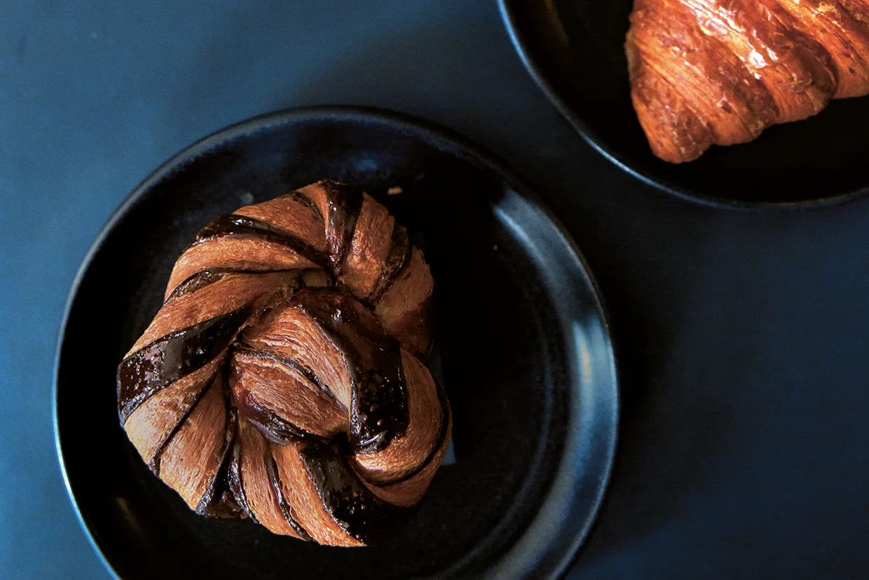 En chokladdoppad bakelse ligger på en svart tallrik intill kanten av en annan tallrik med en croissant, allt mot en mörk bakgrund.