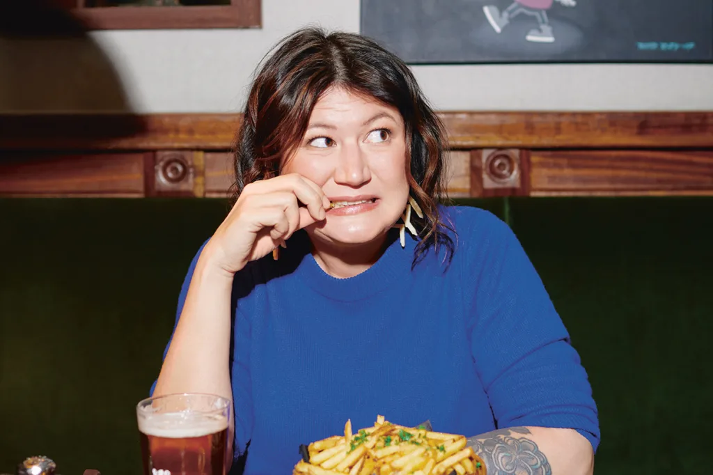 Ina Lundström i blå skjorta sitter i ett bås med ett nervöst ansiktsuttryck och biter på en pommes frites. Framför henne står en tallrik pommes frites och ett glas öl. Bakgrunden består av träpaneler och en inramad tavla.