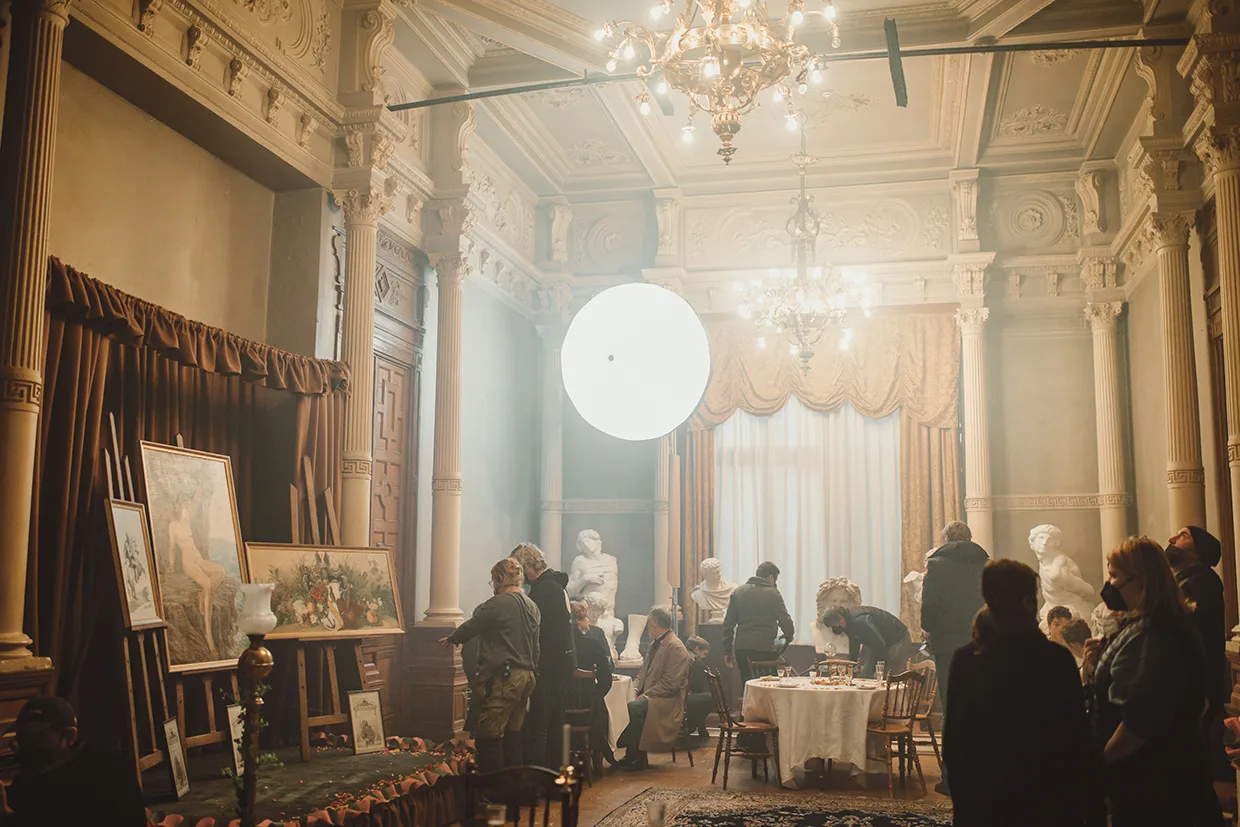 Ett filmteam ställer i ordning en scen i ett utsmyckat, klassiskt rum med pelare, ljuskronor, statyer och draperade gardiner. Människor står runt ett bord och belysningsutrustning syns ovanför.