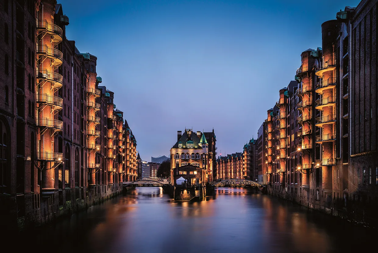 En nattlig vy över det upplysta lagerdistriktet Speicherstadt i Hamburg, Tyskland, med tegelbyggnader som kantar en kanal och en central byggnad på vattnet under en skymningshimmel.
