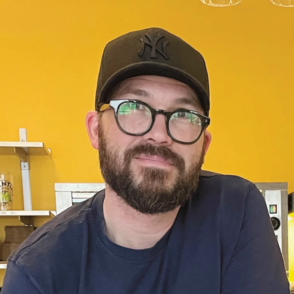 Fredrik Skogdalen med skägg, glasögon och en svart New York Yankees-keps sitter framför en ljusgul vägg, iklädd en mörk T-shirt. Hyllor och köksutrustning syns i bakgrunden.