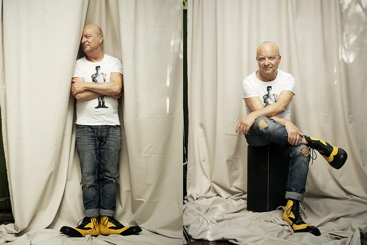 En skallig Jonas Gardell i vit t-shirt, jeans och gulsvarta clownstövlar står med korslagda armar på ena bilden och sitter på en svart låda på den andra och poserar framför beige gardiner.
