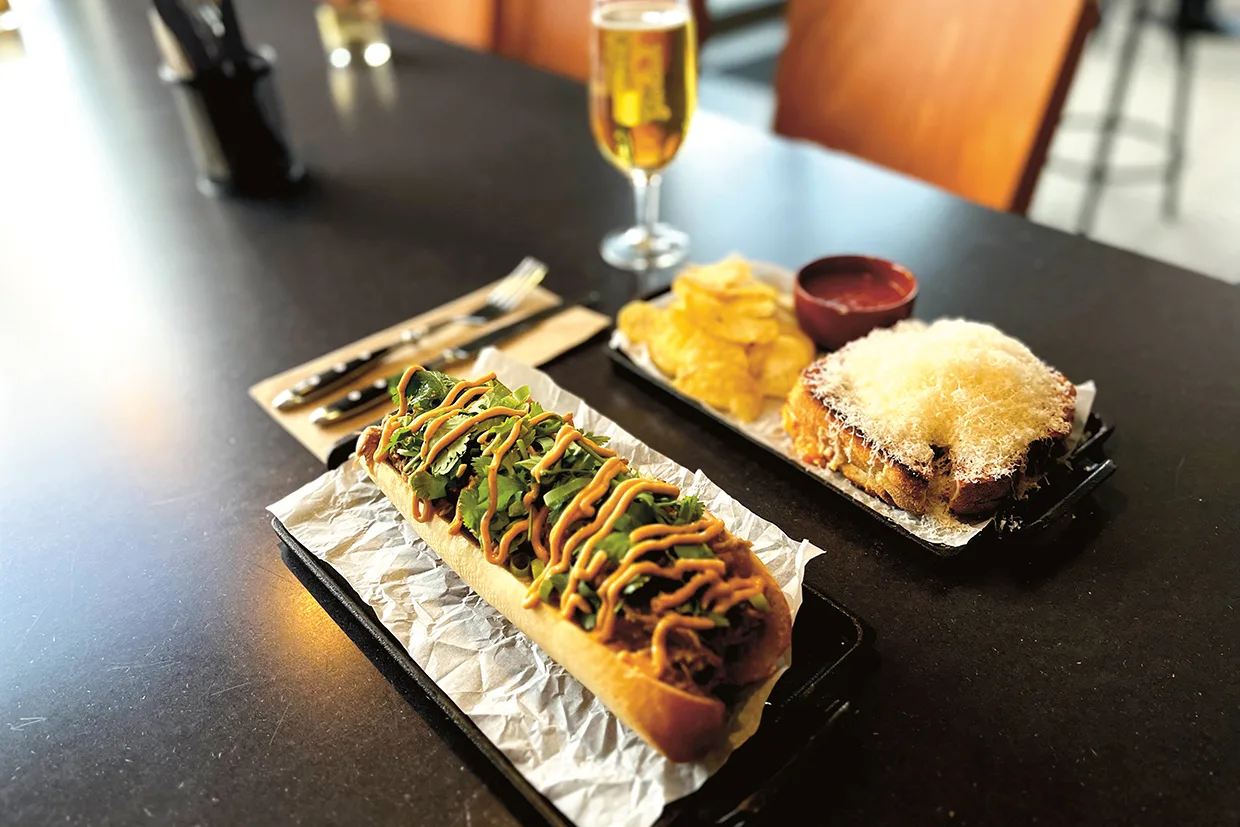 En lång smörgås med grönsaker och sås, en hög med potatischips, en smörgås med riven ost och ett glas öl är placerade på ett mörkt bord i en restaurangmiljö.