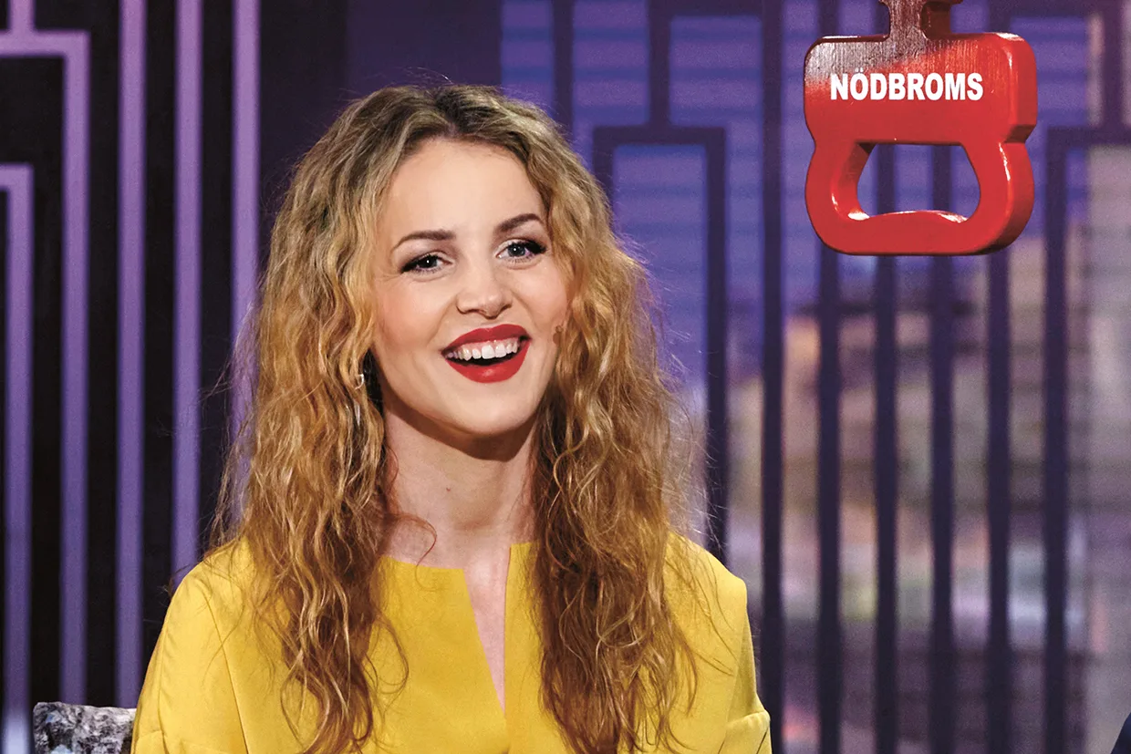 En leende Marie Agerhäll med långt, lockigt blont hår och gul skjorta sitter inomhus. I bakgrunden syns ett stort rött nödbromshandtag märkt NODBROMS. Miljön ser ut att vara en studio eller en talkshow.