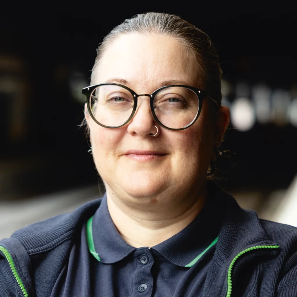 Konduktör Siv Rönnholm med ljus hy, glasögon och en ring i näsan ler mjukt. Hen bär en marinblå skjorta med grön krage och en mörk kavaj och har håret bakåtkammat. Bakgrunden är svag och oskarp.