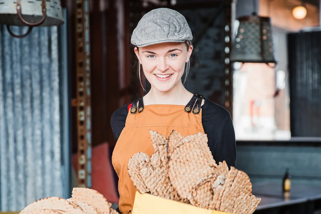 En leende person med grå keps och brunt förkläde håller i en gul låda fylld med stora, runda knäckebröd i en industristil.