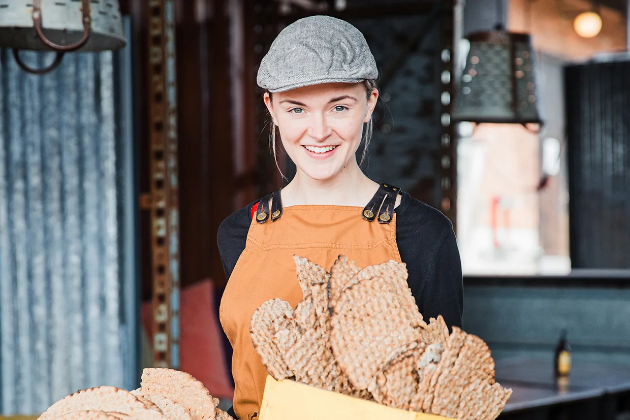 En leende person med grå keps och brunt förkläde håller i en gul låda fylld med stora, runda knäckebröd i en industristil.