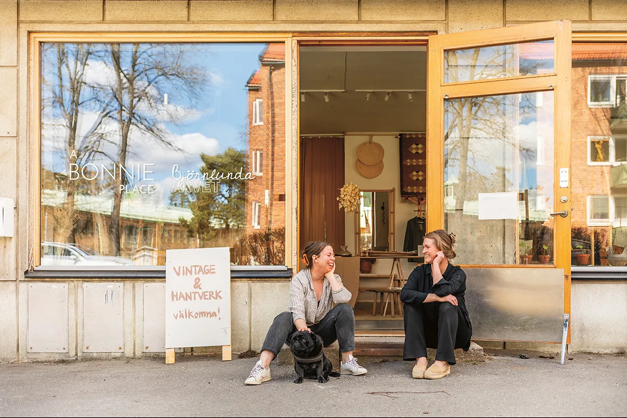 Två kvinnor sitter och pratar utanför ett skyltfönster med stora fönster och en öppen dörr. Mellan dem sitter en svart hund. På en skylt står det Vintage & Hantverk, välkomna. Butikens namn, BONNIE PLACE, syns på fönstret.