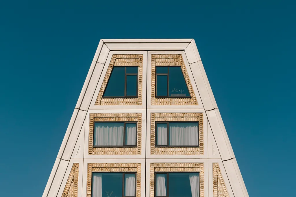 En modern byggnad med triangulärt tak, beige tegelfasad och stora fyrkantiga fönster med svarta ramar mot en klarblå himmel.