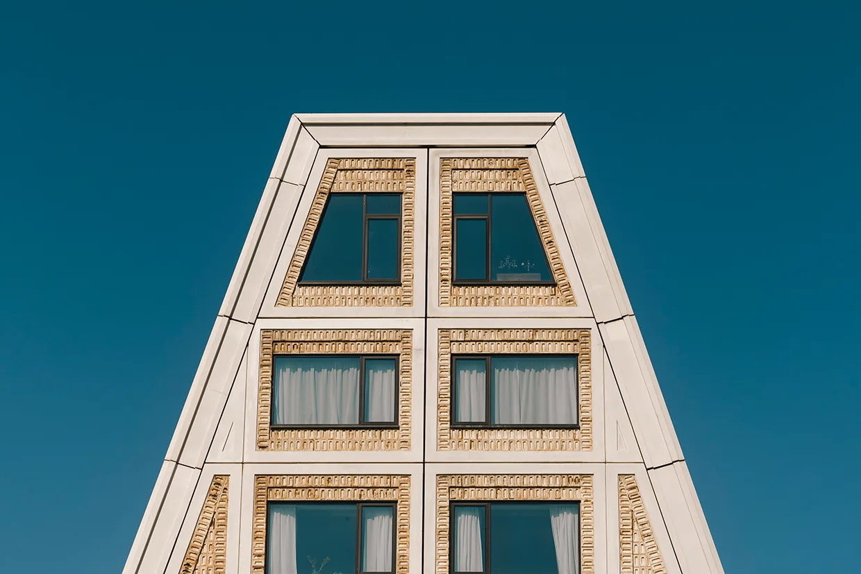 En modern byggnad med triangulärt tak, beige tegelfasad och stora fyrkantiga fönster med svarta ramar mot en klarblå himmel.