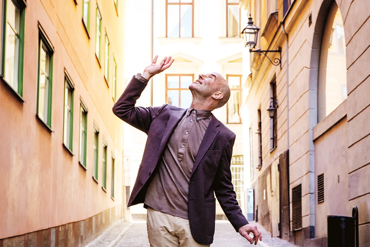 Mark Levengod i kavaj och beige byxor står i en smal, solbelyst gränd och tittar och gestikulerar uppåt med en uppsträckt hand, omgiven av höga, färgglada byggnader med stora fönster.