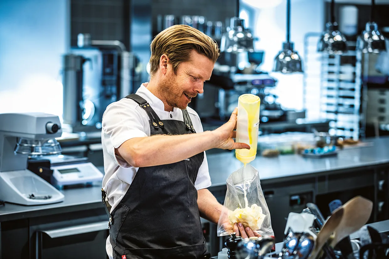 En kock i svart förkläde och vit skjorta pressar ner en gul flaska i en spritspåse fylld med grädde eller smet i ett modernt kök i rostfritt stål.