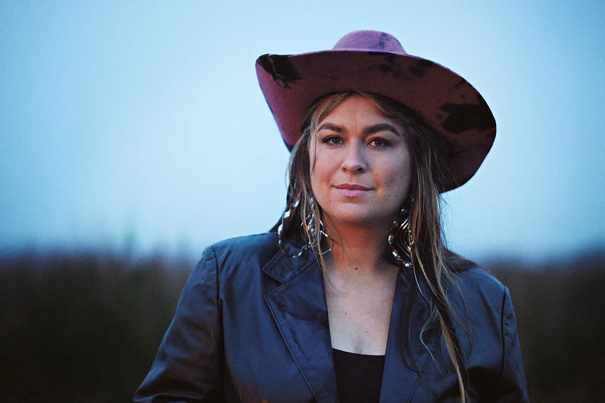 Linnea Henriksson med en stor rosa och svart cowboyhatt, en mörk jacka och dinglande örhängen står utomhus med en suddig bakgrund och blå himmel.