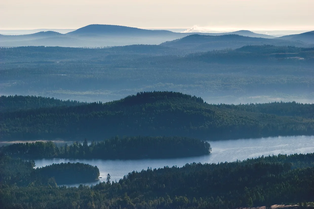 En naturskön vy över trädklädda kullar och öar med en slingrande sjö, allt inbäddat i mjuk dimma och blåtonat ljus under en disig himmel.