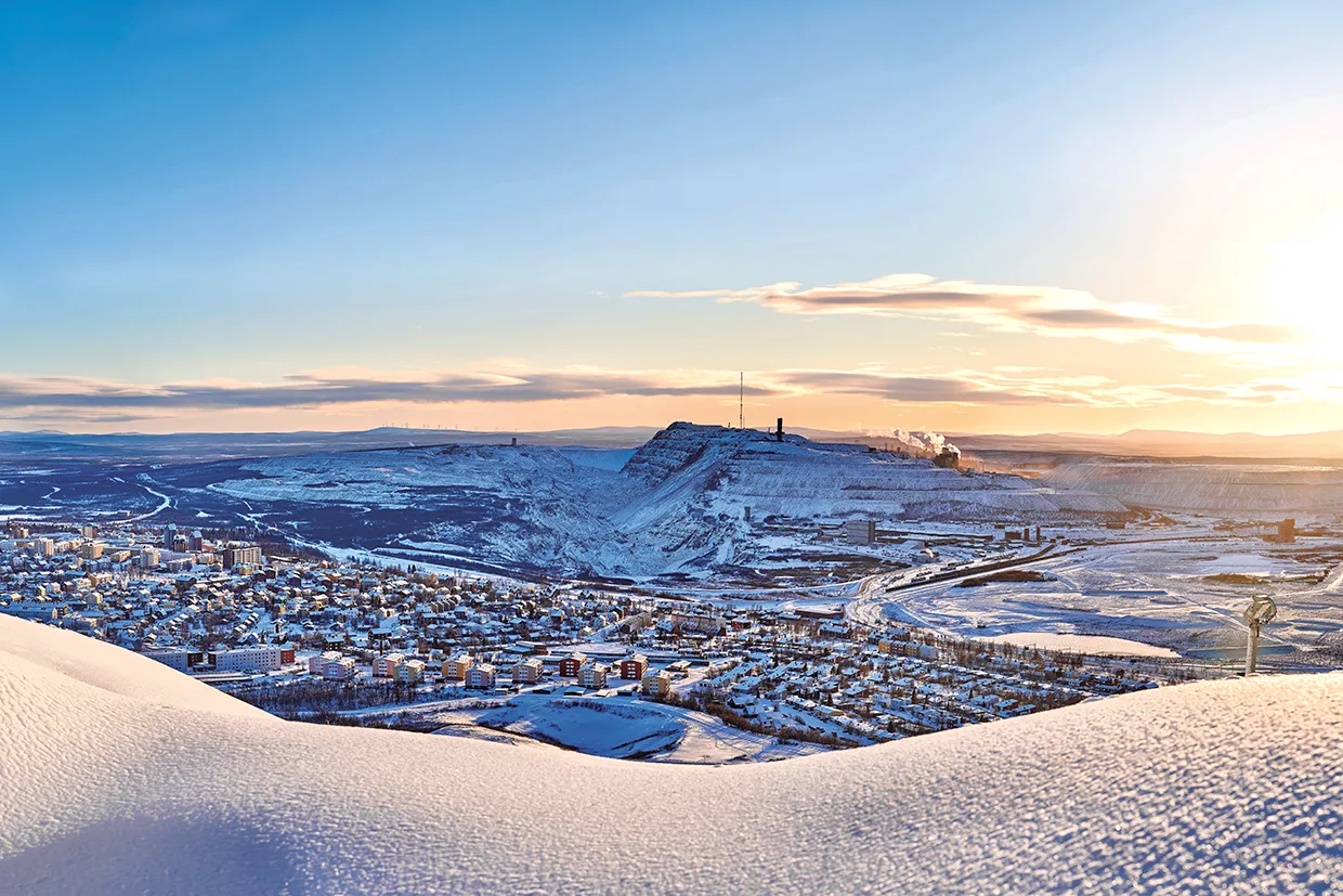 En snöig stad ligger vid foten av en kulle med industribyggnader och rök i fjärran, under en ljus, klar himmel vid soluppgång eller solnedgång. Snön täcker landskapet och reflekterar det gyllene solljuset.