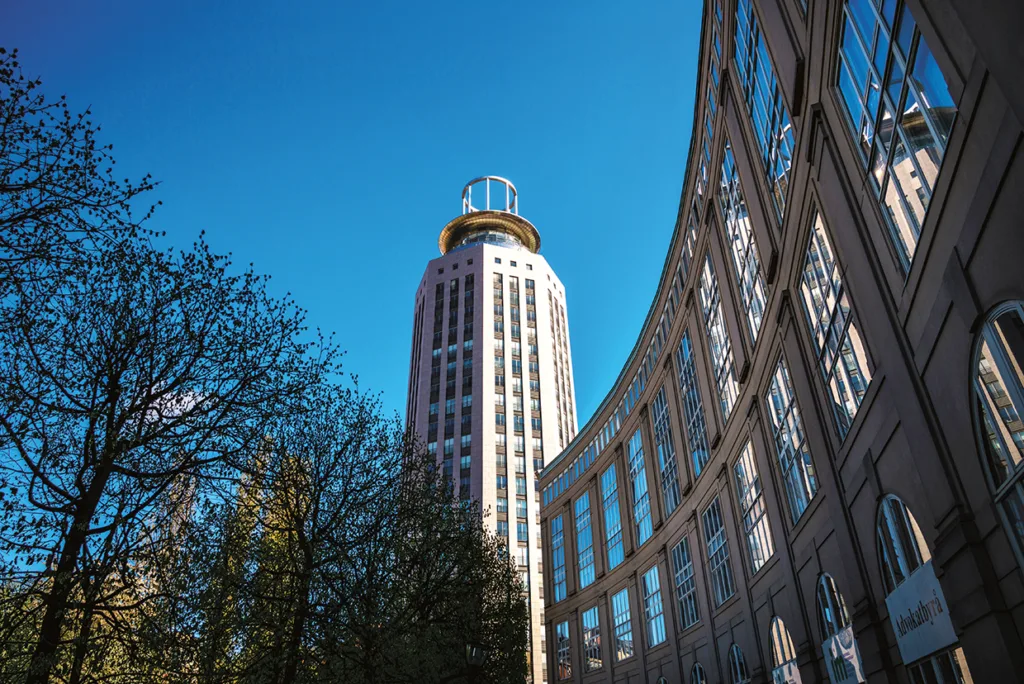 En hög, cylindrisk byggnad med en cirkulär struktur på toppen står bredvid en böjd byggnad med glasfasad under en klarblå himmel, med träd synliga i förgrunden.