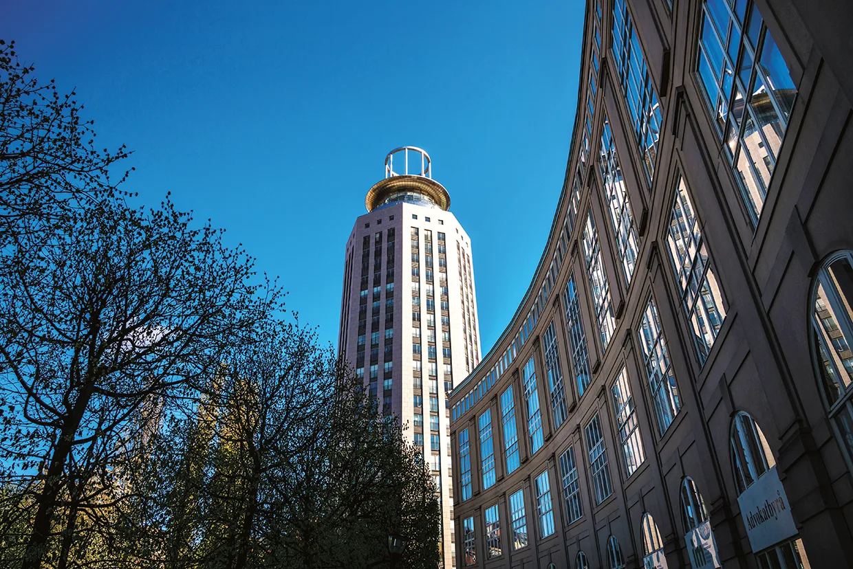 En hög, cylindrisk byggnad med en cirkulär struktur på toppen står bredvid en böjd byggnad med glasfasad under en klarblå himmel, med träd synliga i förgrunden.