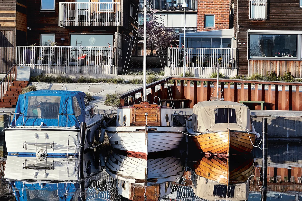 Tre små båtar ligger sida vid sida i lugnt vatten och speglar sina bilder. Bakom dem ligger moderna byggnader vid vattnet med balkonger, stora fönster och träfasader.