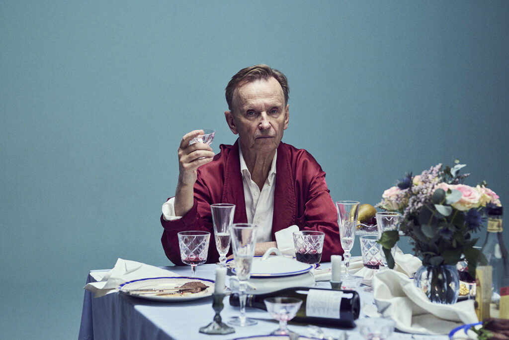 En man i röd kavaj sitter vid ett stökigt bord med tomma vinglas, tallrikar och blommor, håller upp ett glas och tittar rakt in i kameran mot en blå bakgrund.