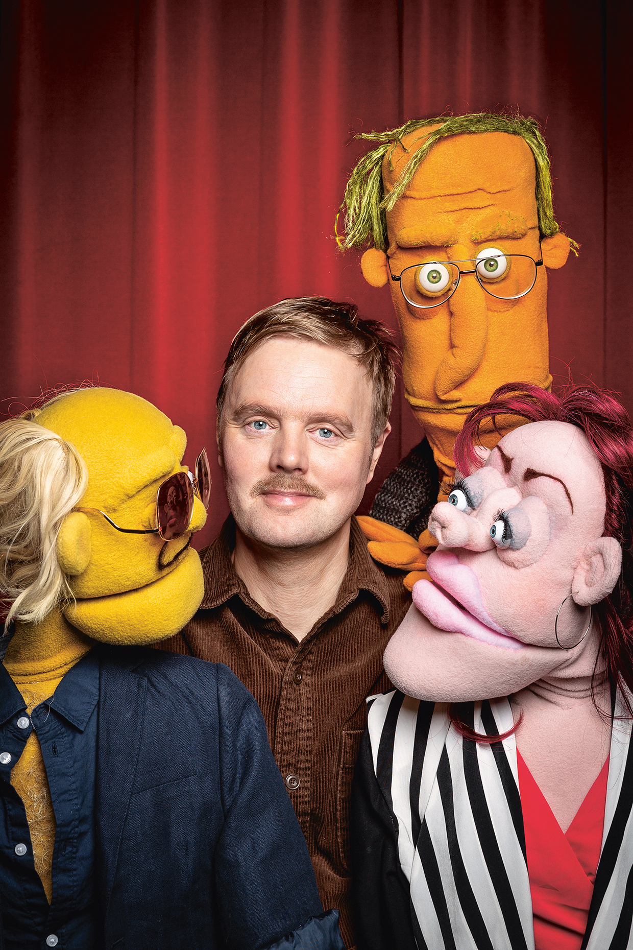 En man med blont hår och mustasch står framför en röd gardin, omgiven av tre färgglada dockor med uttrycksfulla ansikten, som alla tittar mot honom.