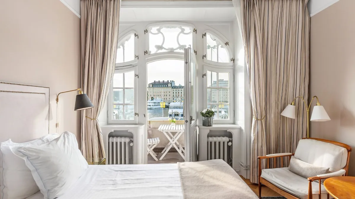 Elegant sovrum med en prydligt bäddad säng, fåtölj och höga fönster med utsmyckade lister som leder ut till en balkong med utsikt över en stad vid vattnet. Ljusa gardiner ramar in fönstren och det finns ett litet bord med stolar utanför.