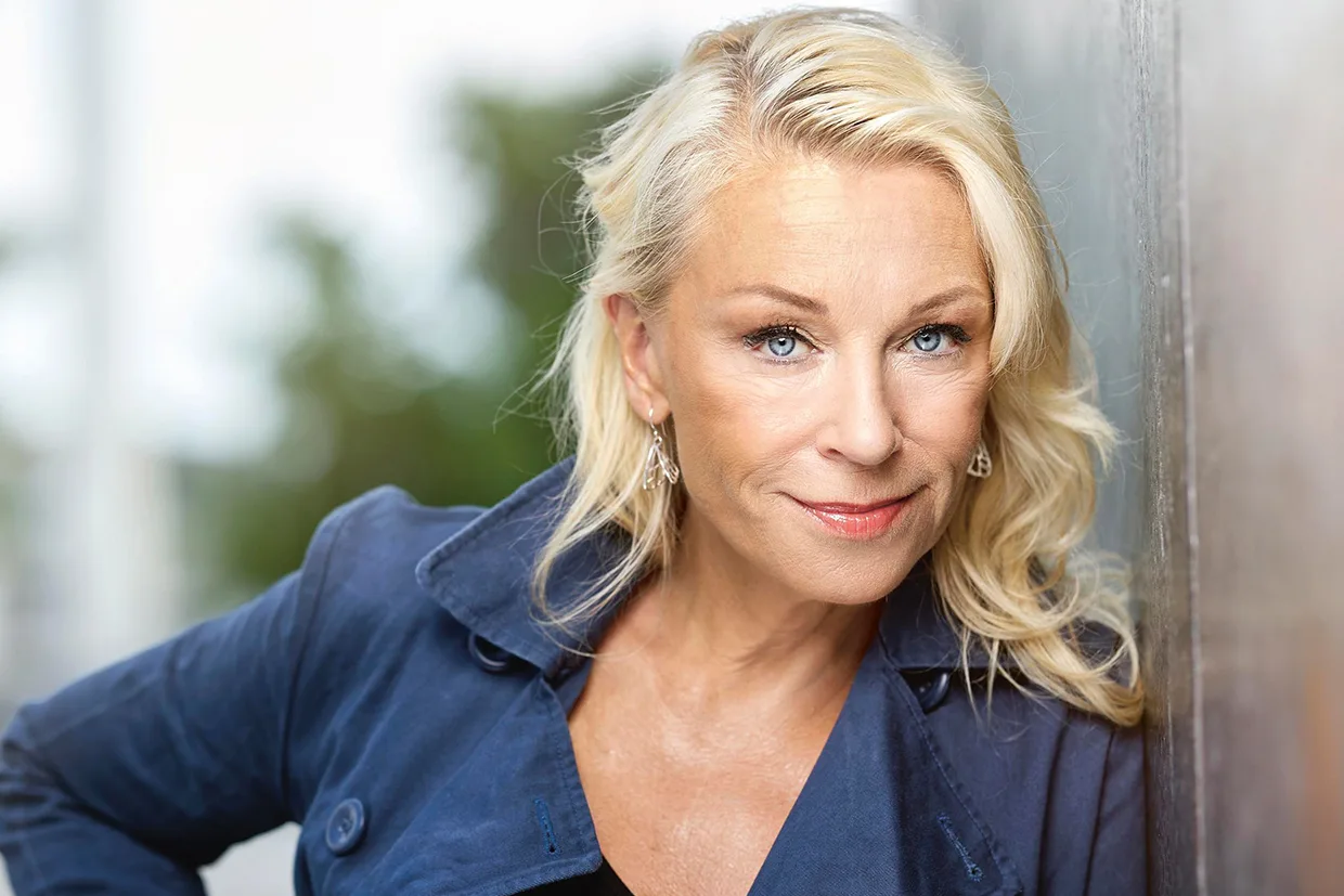 Katarina Ewerlöf med långt blont hår och blå ögon, klädd i en marinblå jacka, lutar sig mot en vägg utomhus och ler mjukt mot kameran. Bakgrunden är suddig med grönska.
